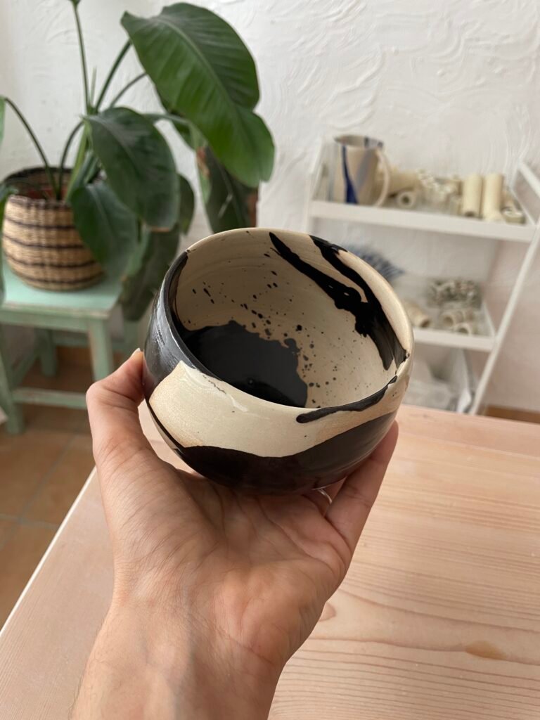 Hand hält eine handgefertigte Keramikschale in Creme und Schwarz, innen und außen mit expressiven, schwarzglänzenden Glasurverläufen. In einem hellen Atelierraum mit Pflanzen und Regalen.